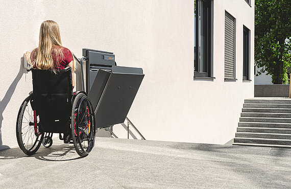 Eine Rollstuhlfahrerin öffnet die Plattform mithilfe der an der Wand montierten Rufstelle.